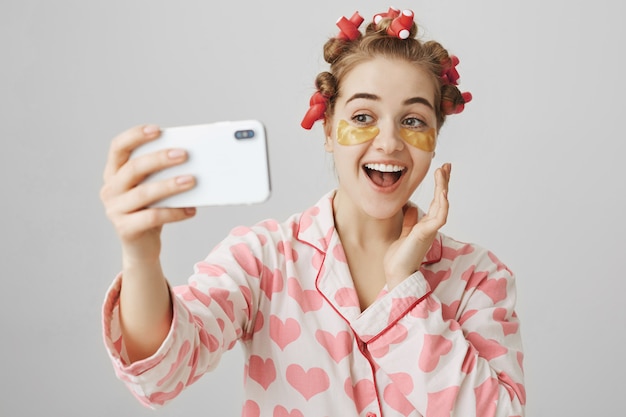 Free photo cheerful smiling girl in hair curlers and eye patches taking selfie wearing nightwear