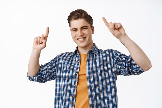 Cheerful smiling guy pointing fingers up and looking happy, showing promo, logo or banner above head, recommend something, click on link, standing over white background
