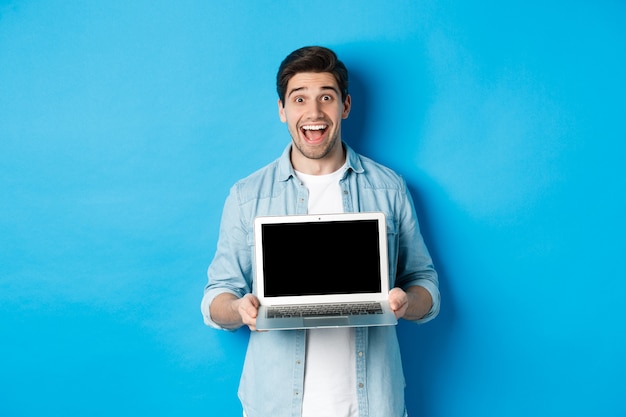 쾌활한 웃는 남자가 프레젠테이션을 하고 노트북 화면을 보여주고 행복해 보이며 파란색 배경 위에 서 있습니다.
