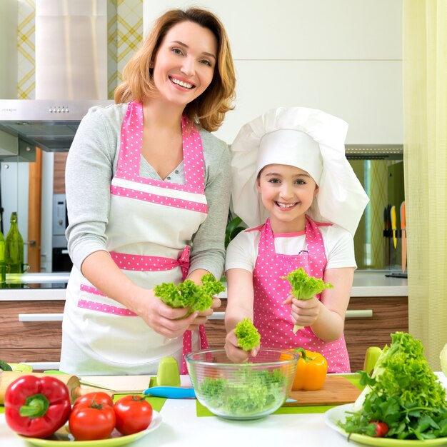 Cheerful smiling mother and daughter cooking a salad at the kitchen.