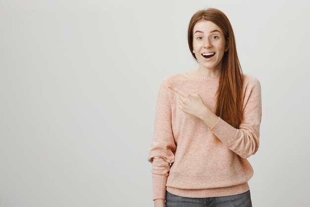 Cheerful smiling redhead girl pointing finger left