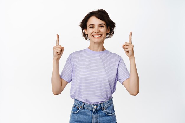 Cheerful smiling woman pointing fingers up, showing advertisement, empty space for logo, company service info, standing against white background.