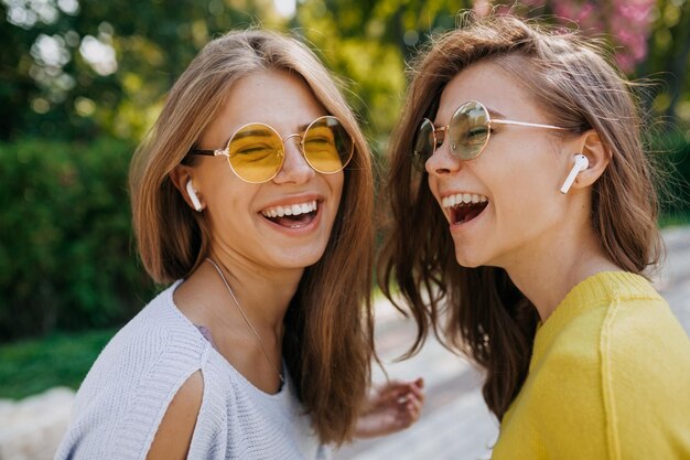 Веселая улыбающаяся женщина с широкой улыбкой смеется с закрытыми глазами во время прослушивания музыки с сестрой Фото двух эмоциональных подруг на улице