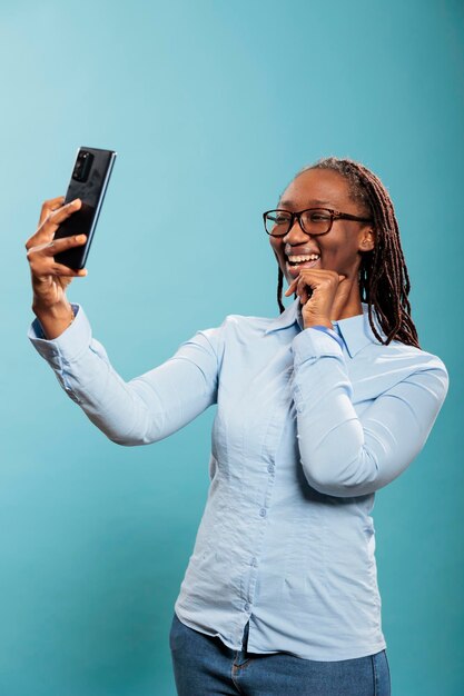 ソーシャルメディアの写真を撮っている間、携帯電話のカメラで笑っている陽気な笑顔の若い大人の人。自撮り写真を撮る現代のタッチスクリーンスマートフォンで陽気な自信のある女性。