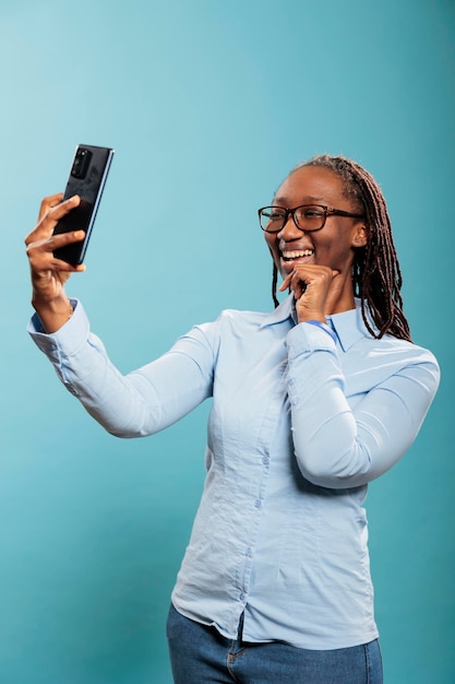 Cheerful smiling young adult person smiling at phone camera while taking picture for social media. Cheerful confident woman with modern touchscreen smartphone taking selfie photo.