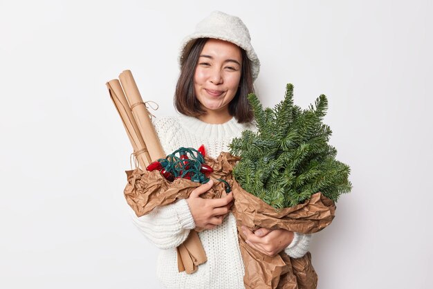 陽気な笑顔の若いブルネットのアジアの女性は暖かいニットのゆるいセーターを着て、パナマは花束と白いスタジオの壁に隔離された装飾のためのアイテムに配置された緑のモミの木の枝を保持します