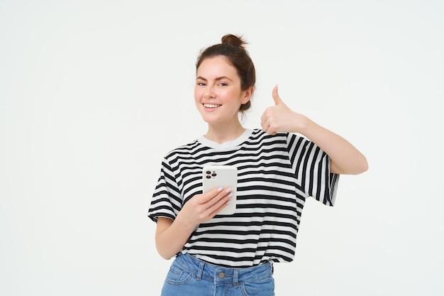 Foto gratuita una giovane donna sorridente e allegra alza il pollice, approva e raccomanda un'app mobile e tiene in mano uno smartphone.