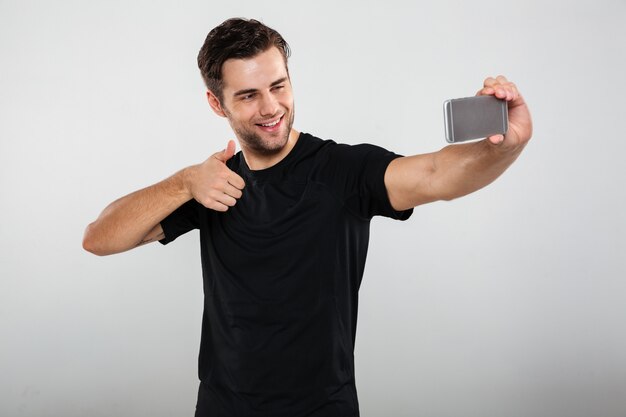 Веселый спортсмен делает selfie по мобильному телефону, показывает палец вверх.