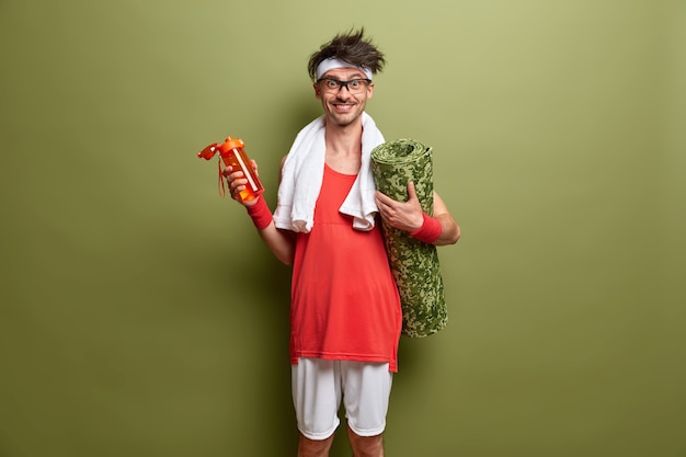 Cheerful sporty man with karemat and bottle of water, going to have physical exercises, being full of energy, enjoys regular training, stands against green wall. Fitness and health concept