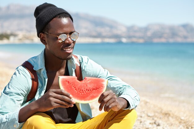 Cheerful student sitting cross-legged on pebble beach and eating fresh juice watermelon