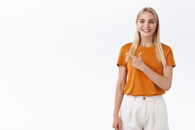 Cheerful stylish blond caucasian woman in orange tshirt smiling enthusiastic pointing finger left at blank white space giving advice what choose standing white background joyful