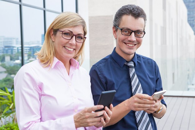 Cheerful successful business people using smartphones and looking at camera in city. 