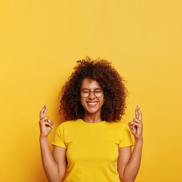 Cheerful superstitious ethnic female hopes for best, crosses finger for good luck, eager to get position in big company, smiles joyfully, wears spectacles and t shirt, isolated on yellow wall.