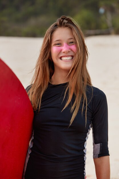 Cheerful surfboarder with toothy smile, dressed in black swimsuit, expresses happiness, has pink zinc sunscreen for protection