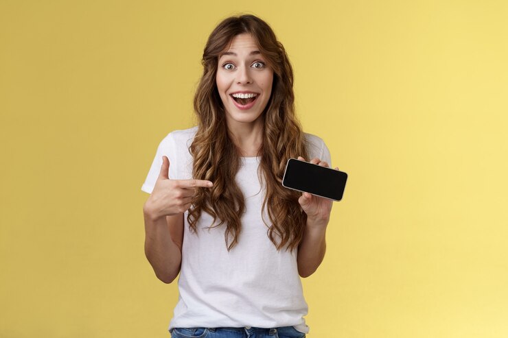 Free Photo cheerful surprised cute lucky girl beat best score awesome game showing smartphone display pointing index finger horizontal screen mobile phone introduce cool app smiling broadly yellow background