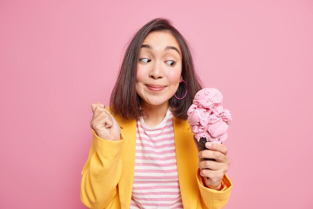 陽気な驚きの若いアジアの女性は、大きな食欲をそそるアイスクリームの笑顔を見て、ピンクの壁に隔離されたファッショナブルな服に身を包んだおいしい休憩ダイエットを楽しんでいます