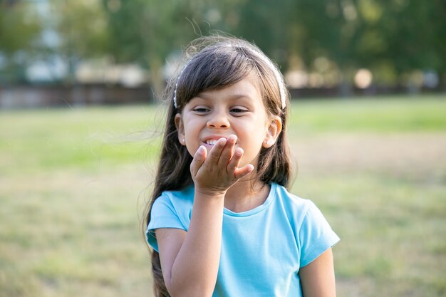 Cheerful sweet black haired girl sending air kiss, posing in park, looking away and smiling. Childhood and outdoor activity concept