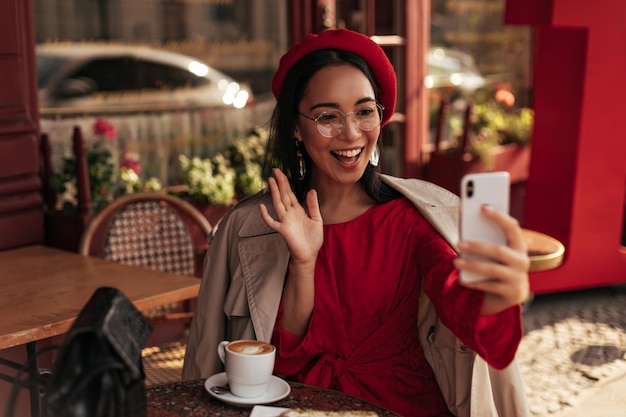 베이지색 트렌치 코트 빨간 드레스와 베레모를 입은 쾌활한 그을린 갈색 머리 아시아 여성이 손을 흔들며 인사하고 휴대폰으로 비디오로 이야기합니다. 매력적인 여성은 거리 카페에 앉아 있습니다.