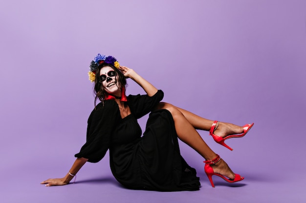 Cheerful tanned model in black dress posing on floor. Flirtatious woman with painted face laughs wholeheartedly.