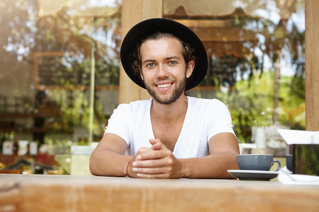 Free photo cheerful tanned student with thick beard having good coffee during lunch, smiling happily, enjoying summer vacations in tropical country