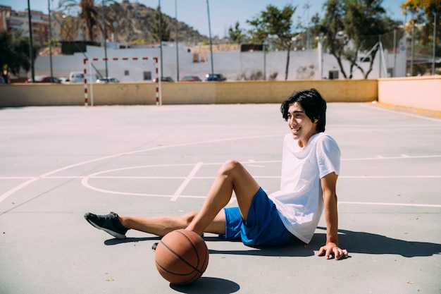 バスケットボールピッチで座っている陽気な十代の少年
