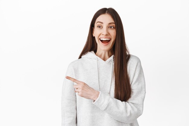 Cheerful teen girl laughing smiling and looking joyful pointing at left side copy space logo showing promotional text standing against white background