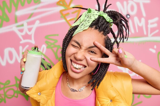 Cheerful teen girl with dreadlocks golden teeth makes peace or victory gesture makes graffiti with aerosol spray dressed in fashionable clothes