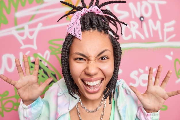 Cheerful teenage girl has fun smiles broadly raises palms looks gladfully at camera glad to make creative graffiti on street wall belongs to youth culture