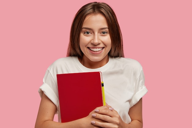 Cheerful teenage girl has toothy smile, healthy skin, long dark hair, carries red notepad with penci