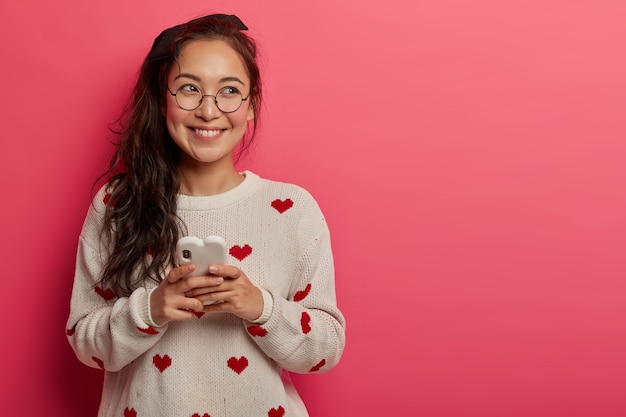 Cheerful teenage girl makes online post via smartphone, looks aside with dreamy expression, updates personal profile, has upbeat mood, wears transparent glasses, casual jumper, stands indoor