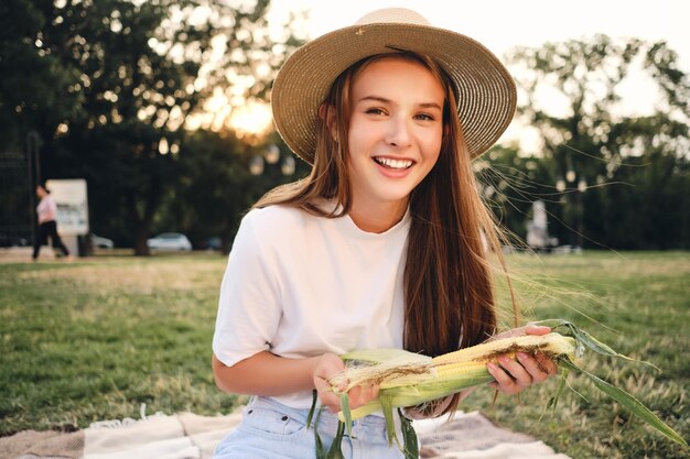 밀짚 모자를 쓴 쾌활한 10대 소녀가 옥수수를 손에 들고 도시 공원에서 피크닉을 할 때 카메라를 행복하게 바라보고 있습니다.