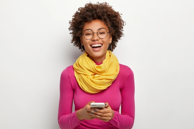 Cheerful teenager with toothy smile, Afro hairstyle, holds modern cell phone, chats online with boyfriend