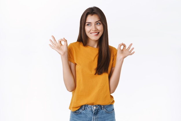 Cheerful, tempting young brunette woman, biting lip with excited, longing face, smiling want something, showing okay ok sign as rating tasty food, standing white background delighted and dreamy