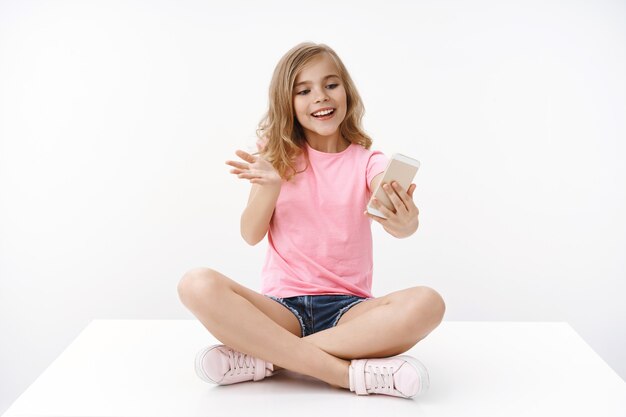 Cheerful tender blond lively enthusiastic young teenage girl, sit crossed legs, hold smartphone, record video-blog become blogger, gesturing excited explain story talking friend, white wall