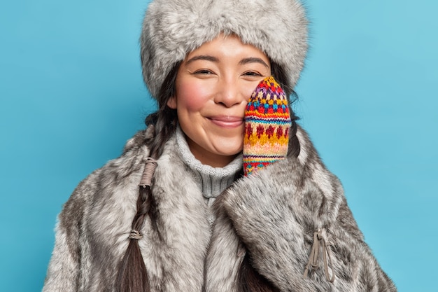 Веселая нежная эскимосская женщина в зимней одежде держит руку на щеке, наслаждается зимними улыбками, с удовольствием позирует на фоне синей стены. Холодный климат
