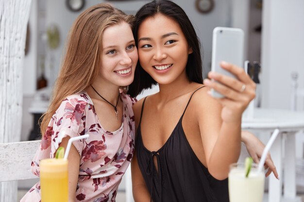 Cheerful two mixed race female friends, make photo in front of smart phone, recreat together in cafeteria, drink cocktails, pose for selfie.