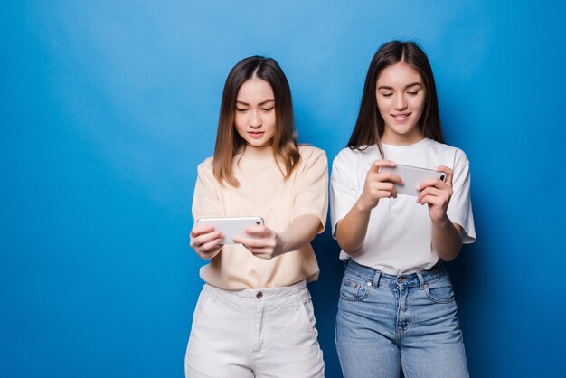 Cheerful two women are playing games with phones in their hands on a blue wall