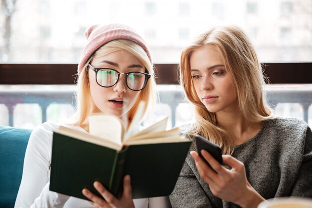 Cheerful two women friends sitting in cafe and reading book