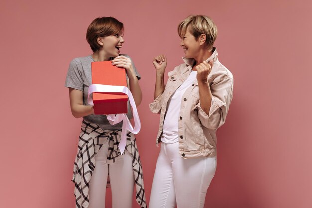 Cheerful two women with short stylish hairstyle in white pants looking each other and posing with oppened red gift box on pink backdrop