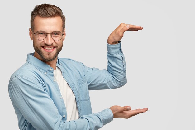 Cheerful unshaven guy stands sideways, holds background, gestures with both hands as if carrying something, shows height of thing