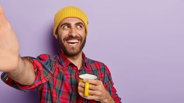 Cheerful unshaven male student enjoys hot drink from yellow mug, takes selfie, keeps arm outstretched, smiles positively, wears yellow hat and checkered shirt