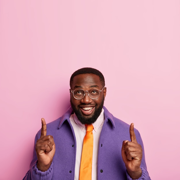 Free photo cheerful unshaven man points both index fingers up, demonstrates successful idea, promots copy space, wears spectacles, orange tie and purple coat