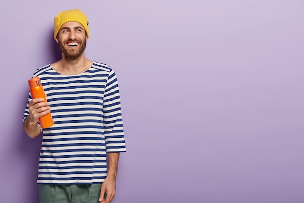 Free photo cheerful unshaven millennial man holds flask with hot beverage, wears yellow hat and striped jumper, being in good mood, stands against purple background, copy space for your promotional content