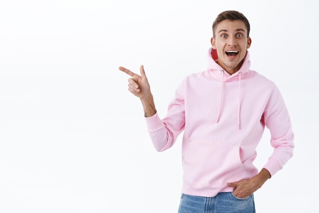 Cheerful upbeat goodlooking blond man wearing pink hoodie react joyful to wonderful news look camera with beaming smile excited showing way pointing finger left white background