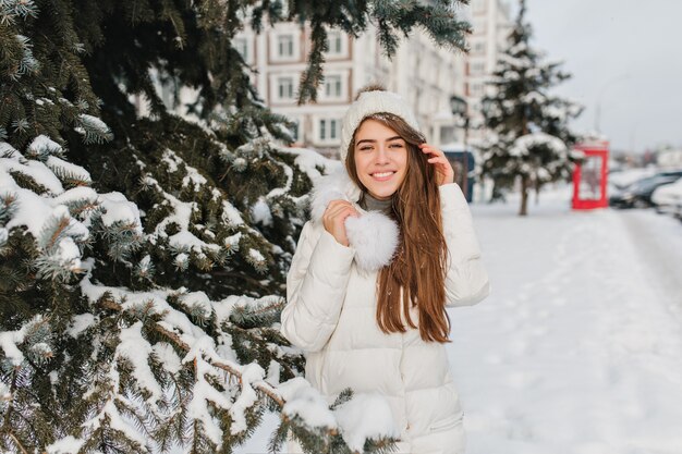 쾌활한 백인 여자는 니트 모자와 따뜻한 코트를 입고 나무 옆에 부드럽게 미소를 지으며 포즈를 취합니다. 겨울 방학 야외 활동을 즐기는 모피 재킷에 긴 머리를 가진 황홀한 여성 모델 ..