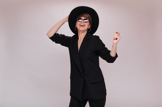 Cheerful woman in black suit and hat dancing on white background. Short-haired lady in dark jacket and pants moves and smiles on isolated