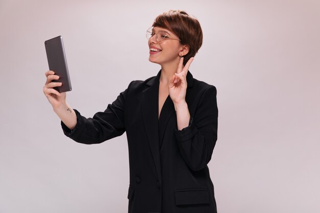 Cheerful woman in black suit holds tablet and takes selfie on isolated background. Happy lady in jacket smiles on white backdrop