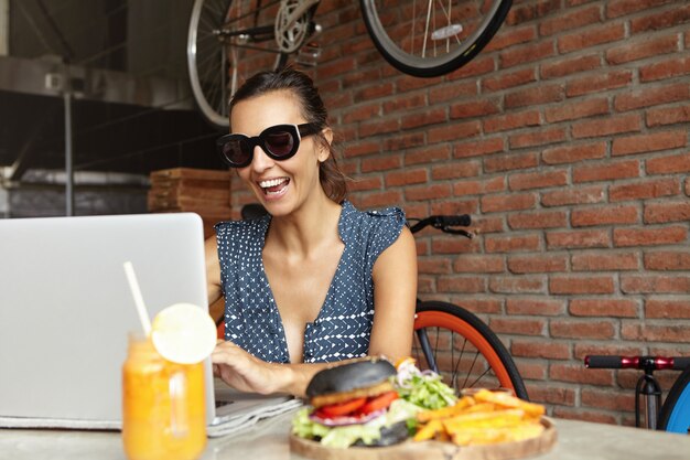 Cheerful woman blogger in trendy sunglasses recording webcam video of herself to post it on her blog, using wireless internet connection