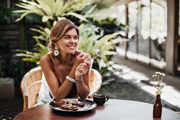 Cheerful woman in brown bra and big white earrings smiles widely and rests in street cafe in summer