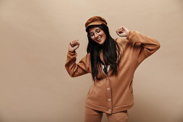 Cheerful woman cashmere sweater and pants moves on isolated Happy brunette girl in cap dances on beige background